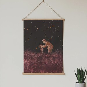 May include: A framed art print depicting a scene of two figures in a field at night, illuminated by glowing orbs. The print is mounted on wooden bars and suspended by a natural cord, creating a minimalist aesthetic. The artwork features warm tones and a dreamy atmosphere.