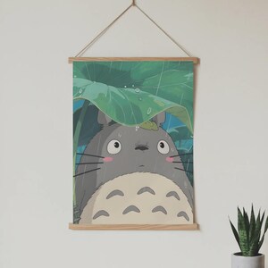 Op de afbeelding: Een ingelijste print met het grijze Totoro-personage uit de animatiefilm My Neighbor Totoro, schuilend onder een groot groen blad in de regen. De print wordt opgehangen door een houten frame en een natuurlijk koord.