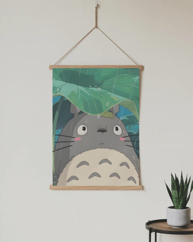 Op de afbeelding: Een ingelijste print met het grijze Totoro-personage uit de animatiefilm My Neighbor Totoro, schuilend onder een groot groen blad in de regen. De print wordt opgehangen door een houten frame en een natuurlijk koord.