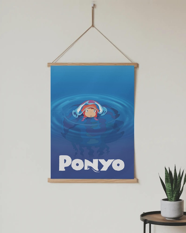 Pu&ograve; includere: Un poster con il personaggio animato Ponyo in una scena oceanica blu. Il poster ha una cornice in legno ed &egrave; sospeso da una corda. La parola "PONYO" &egrave; scritta in lettere bianche in grassetto nella parte inferiore del poster.