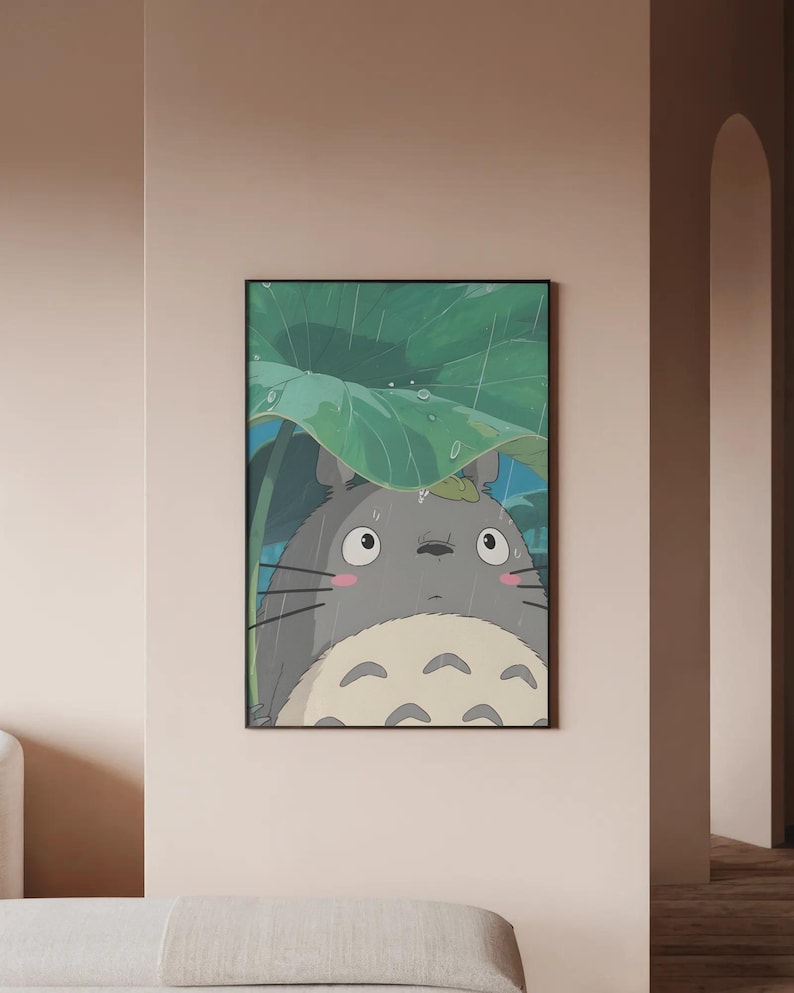 Op de afbeelding: Een ingelijste illustratie van een grijze, pluizige figuur uit de animatiefilm "My Neighbor Totoro", schuilend onder een groot groen blad in de regen. Het kunstwerk heeft een zwarte lijst en een lichtbeige achtergrond.
