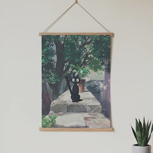 May include: A wall hanging featuring a cartoon illustration of a black cat with large eyes, sitting on a stone wall. The background shows a green tree, a potted plant, and a building. The artwork is framed with wooden bars and suspended by a string.