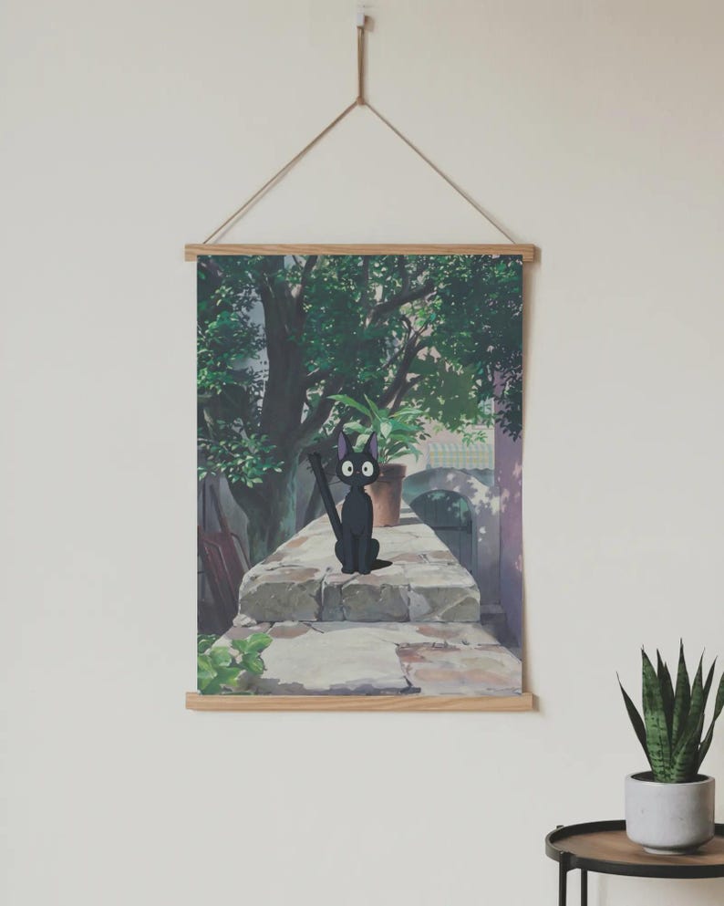 May include: A wall hanging featuring a cartoon illustration of a black cat with large eyes, sitting on a stone wall. The background shows a green tree, a potted plant, and a building. The artwork is framed with wooden bars and suspended by a string.