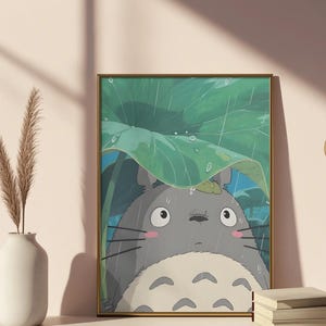 Op de afbeelding: Ingelijste kunstdruk met een grijze Totoro-figuur uit de animatiefilm My Neighbor Totoro, schuilend onder een groot groen blad in de regen. Het kunstwerk heeft een gouden lijst en wordt weergegeven op een wit oppervlak.