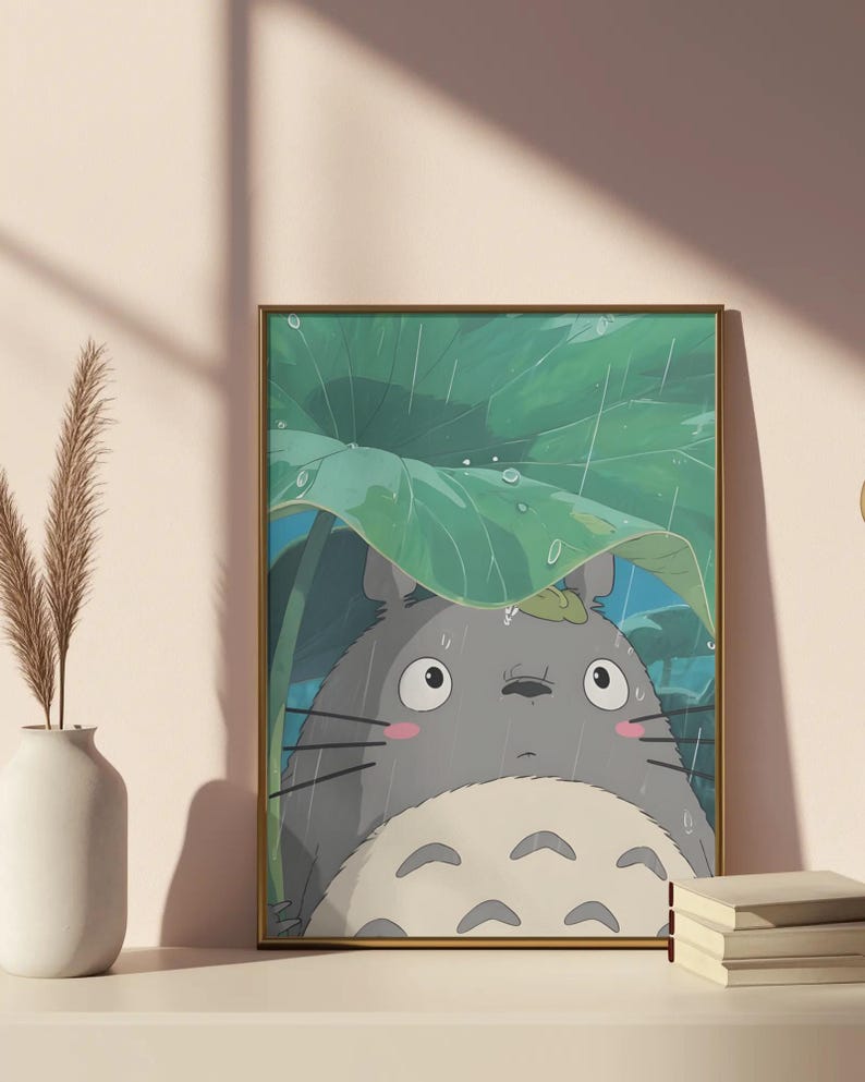Op de afbeelding: Ingelijste kunstdruk met een grijze Totoro-figuur uit de animatiefilm My Neighbor Totoro, schuilend onder een groot groen blad in de regen. Het kunstwerk heeft een gouden lijst en wordt weergegeven op een wit oppervlak.