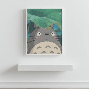 Op de afbeelding: Een ingelijste print met een grijze Totoro-figuur uit de animatiefilm My Neighbor Totoro, schuilend onder een groot groen blad in de regen. Het kunstwerk heeft een witte rand en wordt tentoongesteld op een wit schap.