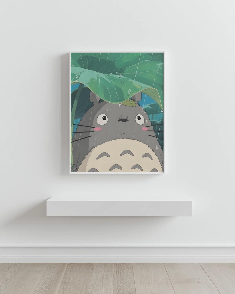 Op de afbeelding: Een ingelijste print met een grijze Totoro-figuur uit de animatiefilm My Neighbor Totoro, schuilend onder een groot groen blad in de regen. Het kunstwerk heeft een witte rand en wordt tentoongesteld op een wit schap.