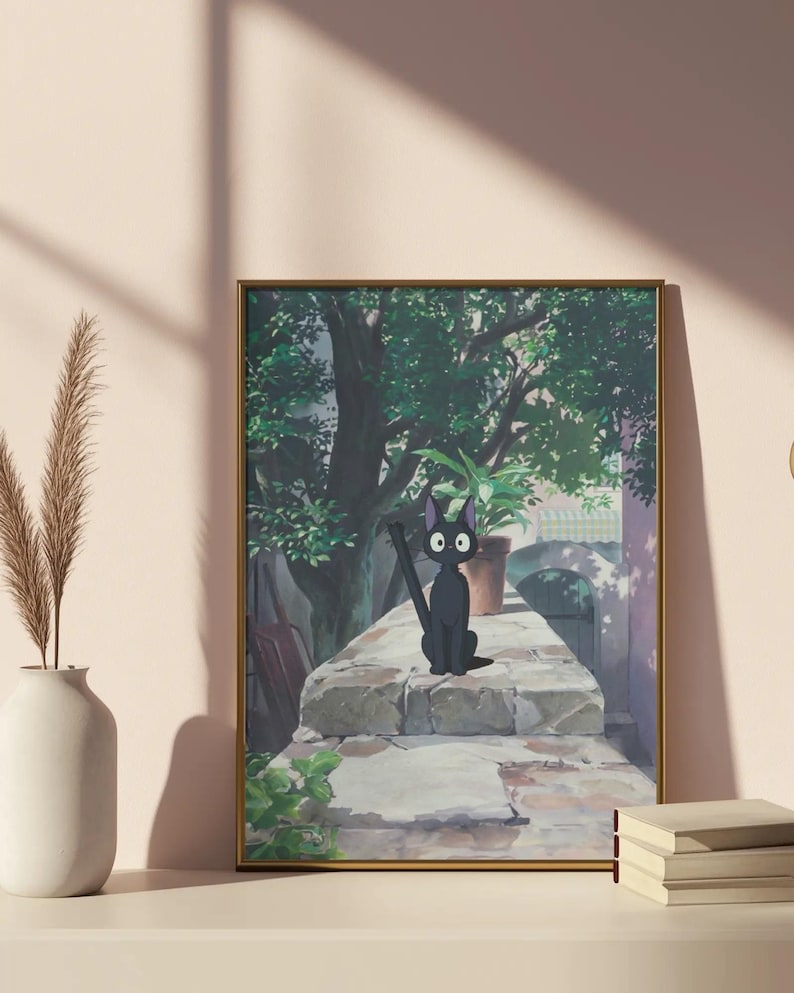 May include: Framed art print featuring a black cat with large eyes, sitting on a stone wall. The background shows a lush green tree, a potted plant, and a glimpse of a building. The artwork is in a gold frame and rests on a white surface.