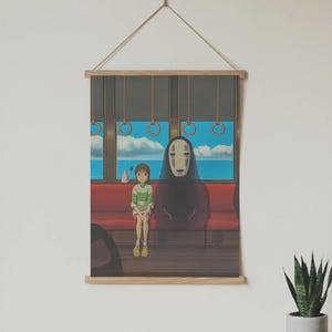 May include: A framed print featuring a scene from an animated film. The artwork depicts two characters on a train, with a scenic ocean view visible through the window. The print is mounted on wooden bars and suspended by a string.