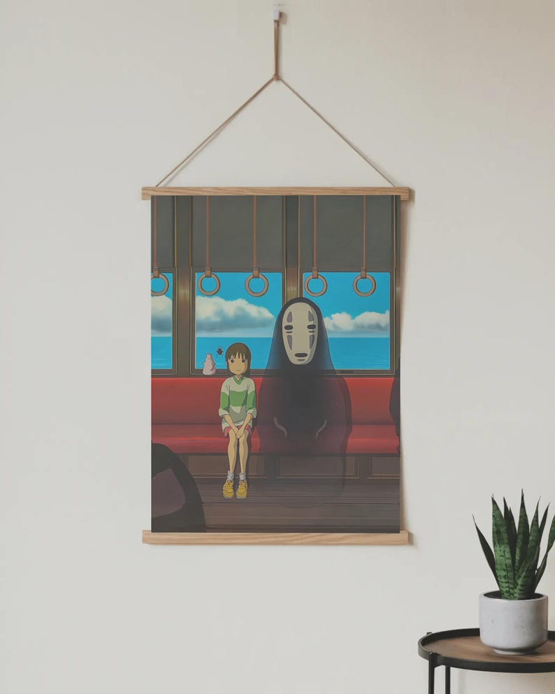 May include: A framed print featuring a scene from an animated film. The artwork depicts two characters on a train, with a scenic ocean view visible through the window. The print is mounted on wooden bars and suspended by a string.