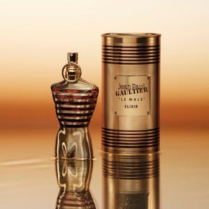 Könnte beinhalten: Ein goldfarbener Parfümflakon und ein passender zylindrischer Behälter. Die Flasche hat die Form eines Torsos mit horizontalen Linien und roten Akzenten. Der Behälter trägt die Aufschrift "Jean Paul Gaultier LE MALE ELIXIR".