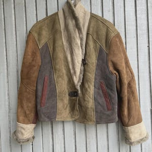 May include: A multi-toned suede jacket with a shearling collar and cuffs. The jacket features a patchwork design in shades of brown, tan, and gray, with a button closure. The jacket is hanging on a metal hanger against a wooden background.