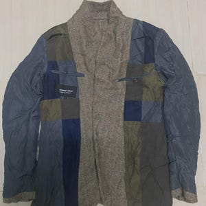 May include: A multi-coloured patchwork jacket featuring panels of blue, green, and grey. The central panel has a tweed-like texture, and the jacket has a high collar. The label "HOMME DEUX" is visible inside.