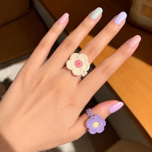 Può includere: Due anelli all'uncinetto a forma di fiore. Un anello presenta un fiore color crema con un centro rosa, indossato sul dito medio. Il secondo anello presenta un fiore viola chiaro con un centro giallo, indossato sul pollice. Entrambi hanno una trama.