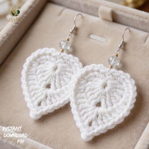 May include: A pair of white, crocheted leaf-shaped earrings. Each earring features a silver-colored hook, a clear bead, and a gold-colored bead. The earrings are displayed in a beige jewelry box. The text "INSTANT DOWNLOAD PDF" is visible.