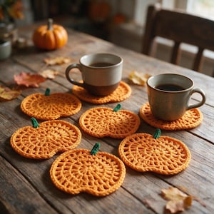 May include: A set of six orange crochet coasters shaped like pumpkins, each featuring a small green stem. Two of the coasters are topped with ceramic mugs filled with dark liquid, arranged on a wooden surface with scattered autumn leaves.