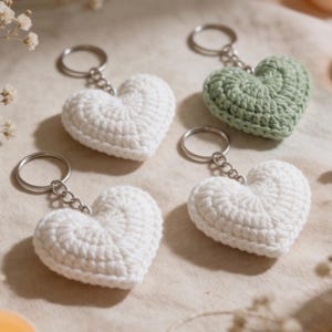 May include: Four crochet heart keychains. Three are white, and one is green. Each heart is attached to a silver-colored keyring. The hearts are made of a textured yarn, and the keychains are arranged on a neutral-toned surface.