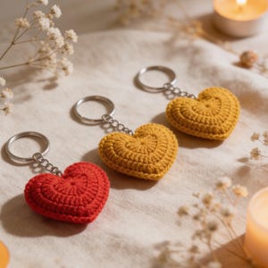 May include: Three crochet heart keychains in red and yellow. Each heart is attached to a silver keyring. The hearts are made with a textured stitch, and the background is a neutral fabric.