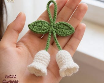 May include: A handmade crochet lily of the valley, featuring two white bell-shaped flowers and green leaves. The design includes the text "instant download pdf". The item is held in a hand.