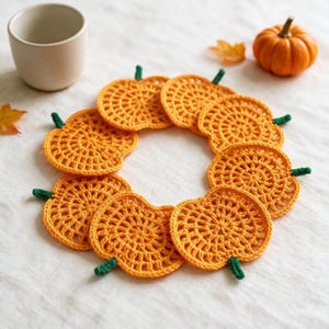 May include: A set of seven orange crochet pumpkin coasters arranged in a circle. Each coaster features a circular orange body with a small green stem attached to the top edge. The crochet pattern consists of radiating stitches creating a textured surface.