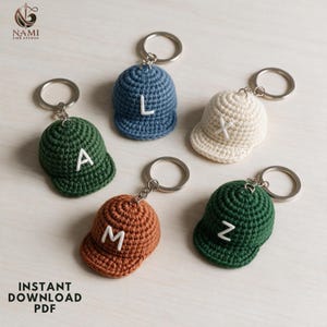 May include: Five crochet baseball cap keychains in blue, cream, brown, and two shades of green. Each cap features a white embroidered letter on the front, including A, L, X, M, and Z. Each item is attached to a silver-toned metal split ring.
