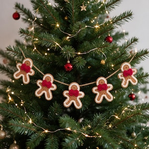 May include: A garland featuring five crocheted gingerbread men figures strung together on a tan twine cord. Each gingerbread man is light brown with white crocheted trim around the edges and a small, textured red bow attached at the neck.
