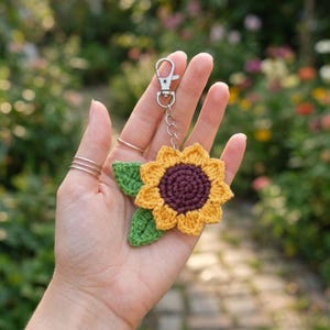 Pode incluir: Um chaveiro de girassol de crochê com uma flor amarela e roxa, folhas verdes e um fecho prateado. O acessório feito à mão é segurado contra um fundo desfocado de vegetação e flores.