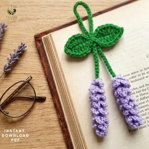 Puede incluir: Un marcapáginas de ganchillo con dos ramitas de lavanda texturizadas en hilo morado claro, unidas por tallos verdes a un par de hojas tejidas y un pequeño lazo en la parte superior, sobre las páginas abiertas de un libro con texto.
