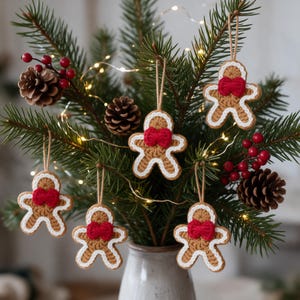 May include: Five crocheted gingerbread man ornaments hanging from a pine tree. Each tan gingerbread figure features a white crocheted outline, a bright red bow at the neck, and a tan string loop for hanging on the branches.