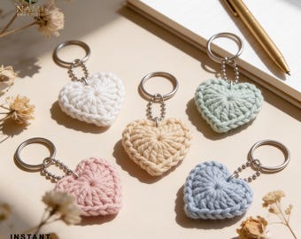 May include: Five heart-shaped keychains made of crocheted yarn in white, beige, light green, light pink, and light blue. Each keychain features a silver-toned metal split ring attached by a small ball chain. The text NAMI EMB STUDIO and INSTANT DOWNLOAD PDF is visible.
