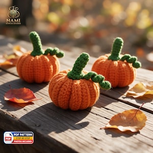 May include: Three small, round, orange crocheted pumpkins with green stems and curly green vines. Each pumpkin features a ribbed texture created by the yarn stitches. The pumpkins are arranged on a wooden surface with scattered autumn leaves.