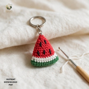 May include: A crochet watermelon slice keychain featuring a red triangular body with black embroidered seeds, a white middle stripe, and a green rind base. The piece is attached to a silver metal split ring, showcasing detailed yarn texture.