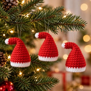 May include: Three small, crocheted Santa hats in bright red with white trim at the base and a white pom-pom at the tip. Each hat is suspended by a thin silver string, featuring a textured yarn construction with a distinct conical shape.
