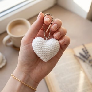 Puede incluir: Un llavero blanco en forma de corazón tejido a ganchillo, sostenido por una mano. El corazón está unido a una cadena plateada. Se ve una pulsera dorada en la muñeca. Una taza de café y un libro están en el fondo.