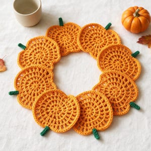 May include: A circular arrangement of eight orange crocheted pumpkin coasters, each featuring a small green stem. The coasters are crafted with a circular stitch pattern and are displayed on a white surface alongside a small ceramic cup and a pumpkin.