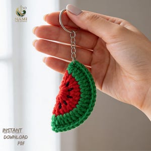 May include: A handmade crochet watermelon slice keychain. The keychain features a red center with black seed details, a green rind, and a silver-colored metal keyring. The text "INSTANT DOWNLOAD PDF" is visible in the lower left corner.