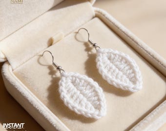 May include: A pair of white, leaf-shaped crochet earrings with silver-colored hooks, displayed in a beige jewelry box. The earrings are handmade with a textured stitch pattern. The text "INSTANT DOWNLOAD PDF" is visible in the lower left corner.
