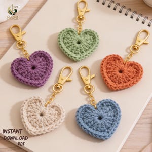 May include: Five crochet heart keychains in various colors: purple, green, orange, cream, and blue. Each heart is attached to a gold-colored clasp and chain. The image also includes the text "INSTANT DOWNLOAD PDF".