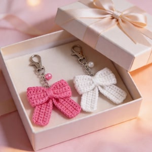 Puede incluir: Dos llaveros de lazo de ganchillo, uno rosa y otro blanco, dentro de una caja de regalo. Cada llavero tiene un cierre plateado y una cuenta decorativa. La caja es blanca con un lazo de cinta de raso en la tapa.