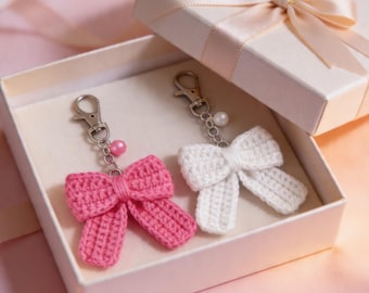 May include: Two crochet bow keychains, one pink and one white, rest inside a gift box. Each keychain has a silver clasp and a decorative bead. The box is white with a satin ribbon bow on the lid.
