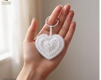 May include: A white crocheted heart keychain featuring a textured yarn pattern attached to a silver-toned metal split ring. The heart is crafted with visible stitches and a small braided loop connecting the heart to the circular metal ring.