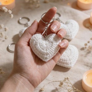 May include: A white, crocheted heart-shaped keychain held in a hand. The keychain has a silver ring and chain. Other white heart keychains are visible in the background, along with lit candles and decorative sprigs.