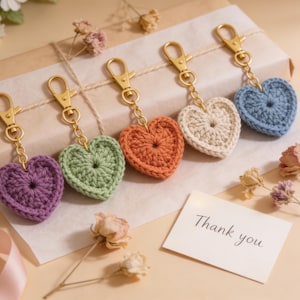 May include: A collection of heart-shaped crochet keychains in various colors: purple, green, orange, cream, and blue. Each keychain has a gold-colored clasp. A small card with "Thank you" is also present.