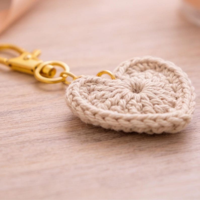 May include: A beige crocheted heart keychain with a gold-colored clasp and ring. The heart is made of a textured yarn and is attached to a gold-toned metal clasp. The background is a light-colored wooden surface.