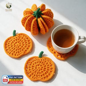 May include: A set of three orange crochet pumpkin items, including one three-dimensional pumpkin and two flat pumpkin-shaped coasters, one of which supports a white ceramic mug filled with tea. The text NAMI EMB STUDIO and PDF PATTERN are visible.