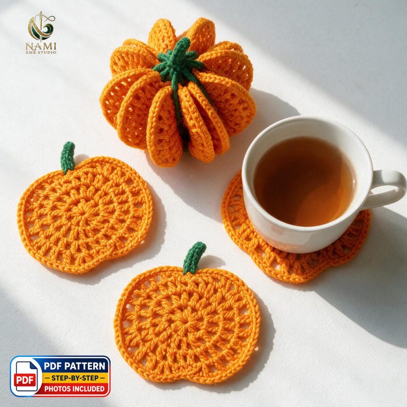 May include: A set of three orange crochet pumpkin items, including one three-dimensional pumpkin and two flat pumpkin-shaped coasters, one of which supports a white ceramic mug filled with tea. The text NAMI EMB STUDIO and PDF PATTERN are visible.