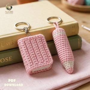 May include: Two crochet keychains featuring a small pink rectangular book and a pink and white pencil. Each item is attached to a silver metal keyring. The book has a textured stitch pattern, and the pencil has a pointed tip. PDF DOWNLOAD text.