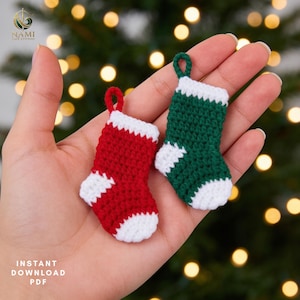 May include: Two miniature crochet stockings, one red and one green, each featuring a white cuff, heel, and toe. Both stockings include a small matching yarn loop at the top for hanging. The text NAMI THE STUDIO and INSTANT DOWNLOAD PDF is visible.