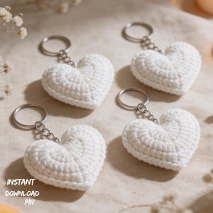 May include: Four white, crocheted heart-shaped keyrings with silver key rings and chains. The keyrings are arranged on a light-coloured surface. The words "INSTANT DOWNLOAD PDF" are printed on the bottom left.