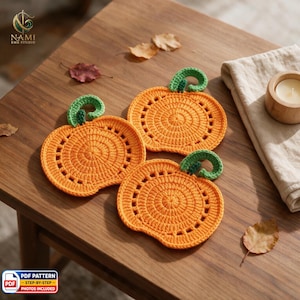 May include: Three orange crochet pumpkin coasters with green stems. Each coaster features a circular center with a decorative openwork border. The pumpkins are arranged on a surface alongside a folded beige cloth and scattered dried autumn leaves.